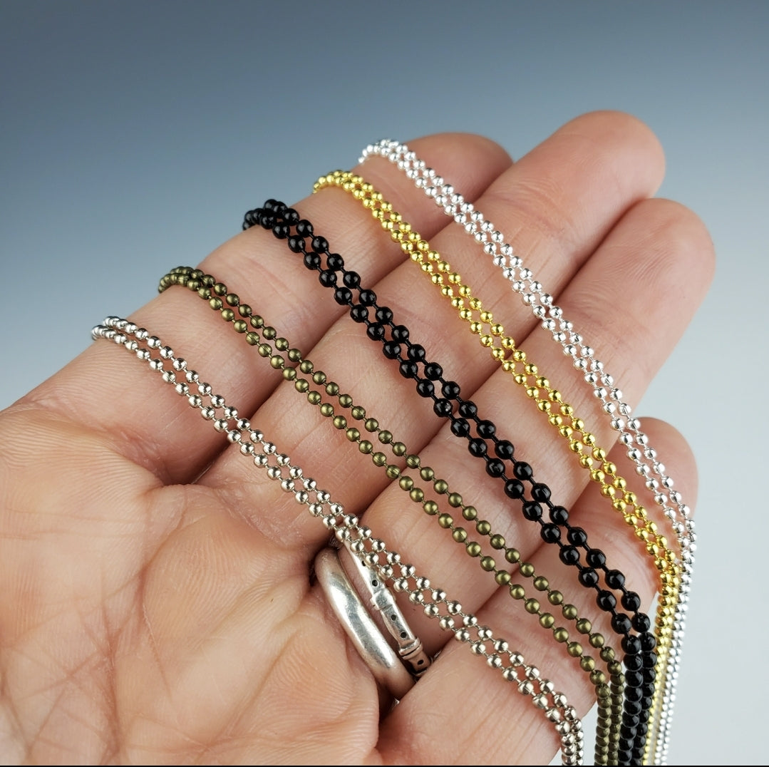 A hand holding five ball chain necklaces in different colors including black, bronze, silver, gold, and white silver.