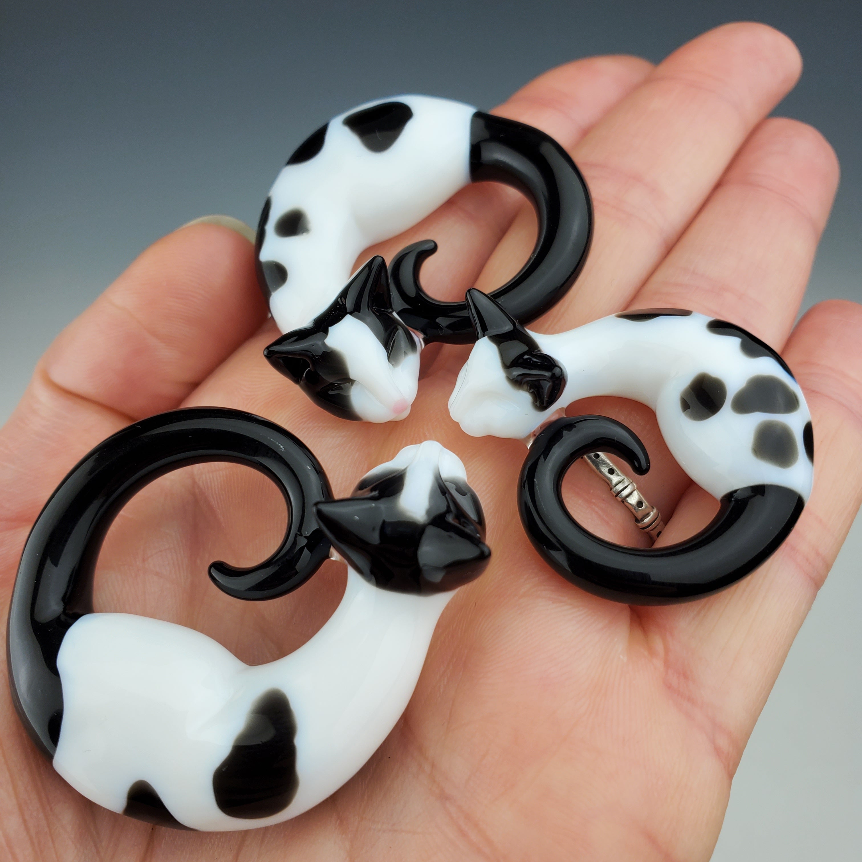 Woman's hand, palm up, holding three small black and white sculpted glass sleeping kitties that can be worn as a necklace or displayed as a memorial.