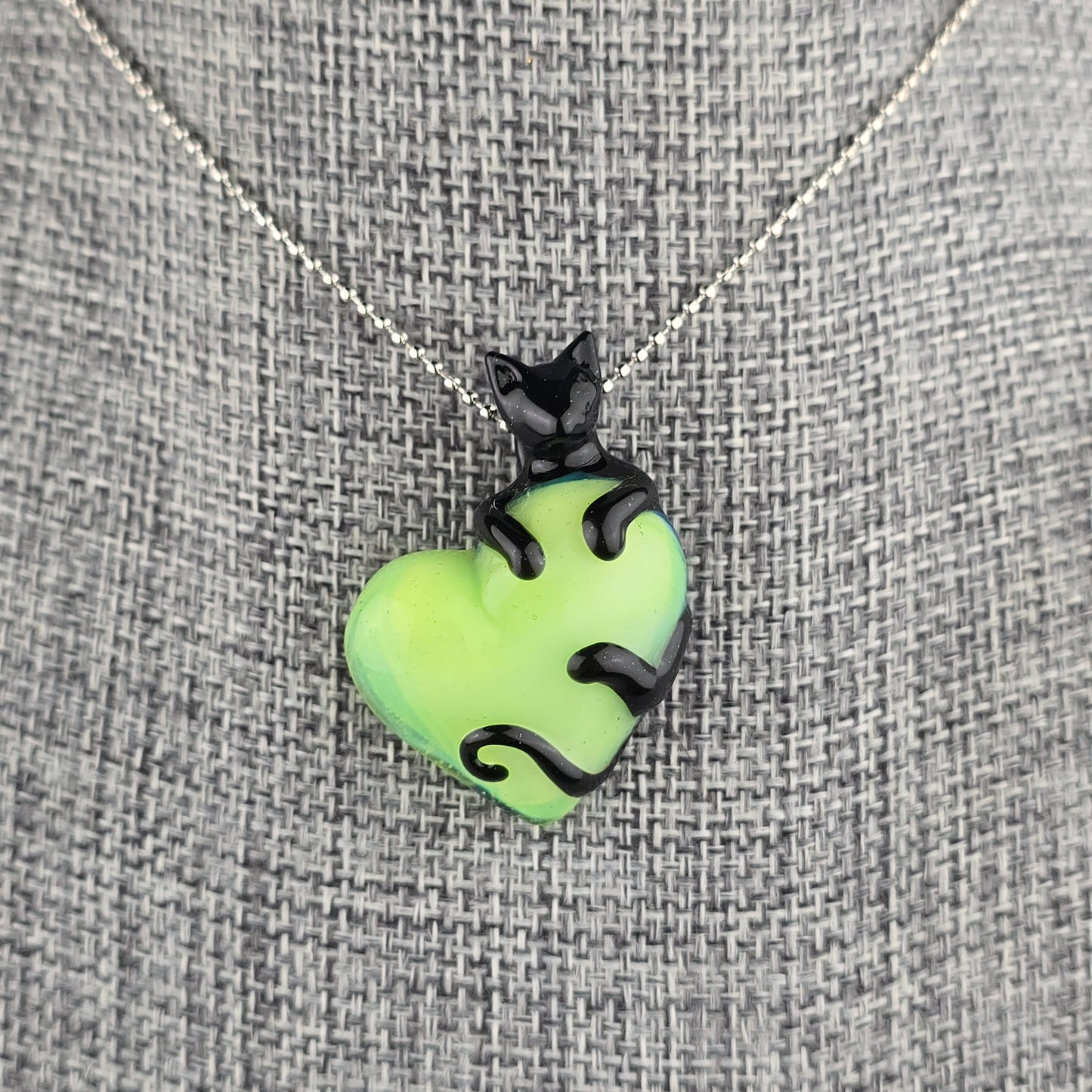 Glass necklace of a sculpted black cat clinging onto a neon green heart