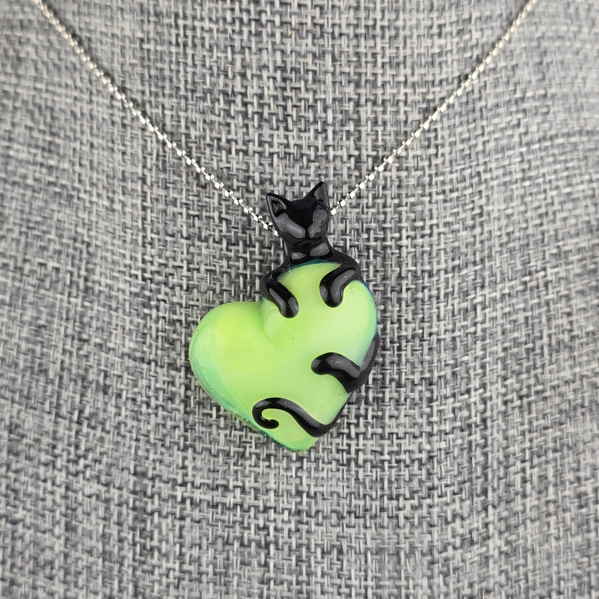 Glass necklace of a sculpted black cat clinging onto a neon green heart