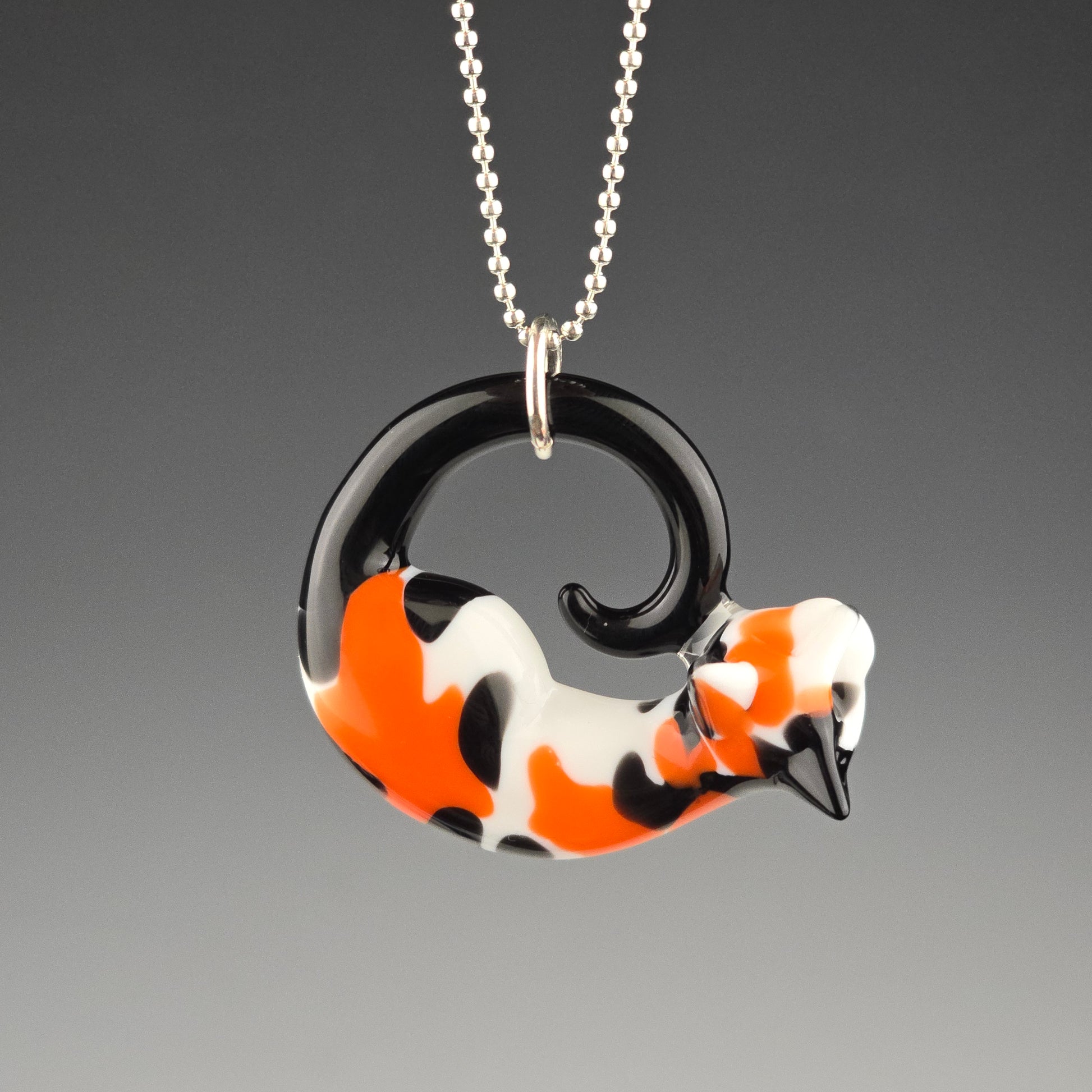 A small glass pendant with a traditional black, orange, and white calico color pattern, shaped like a sleeping kitty. The pendant is attached to a silver ball chain and is displayed against a grey background.