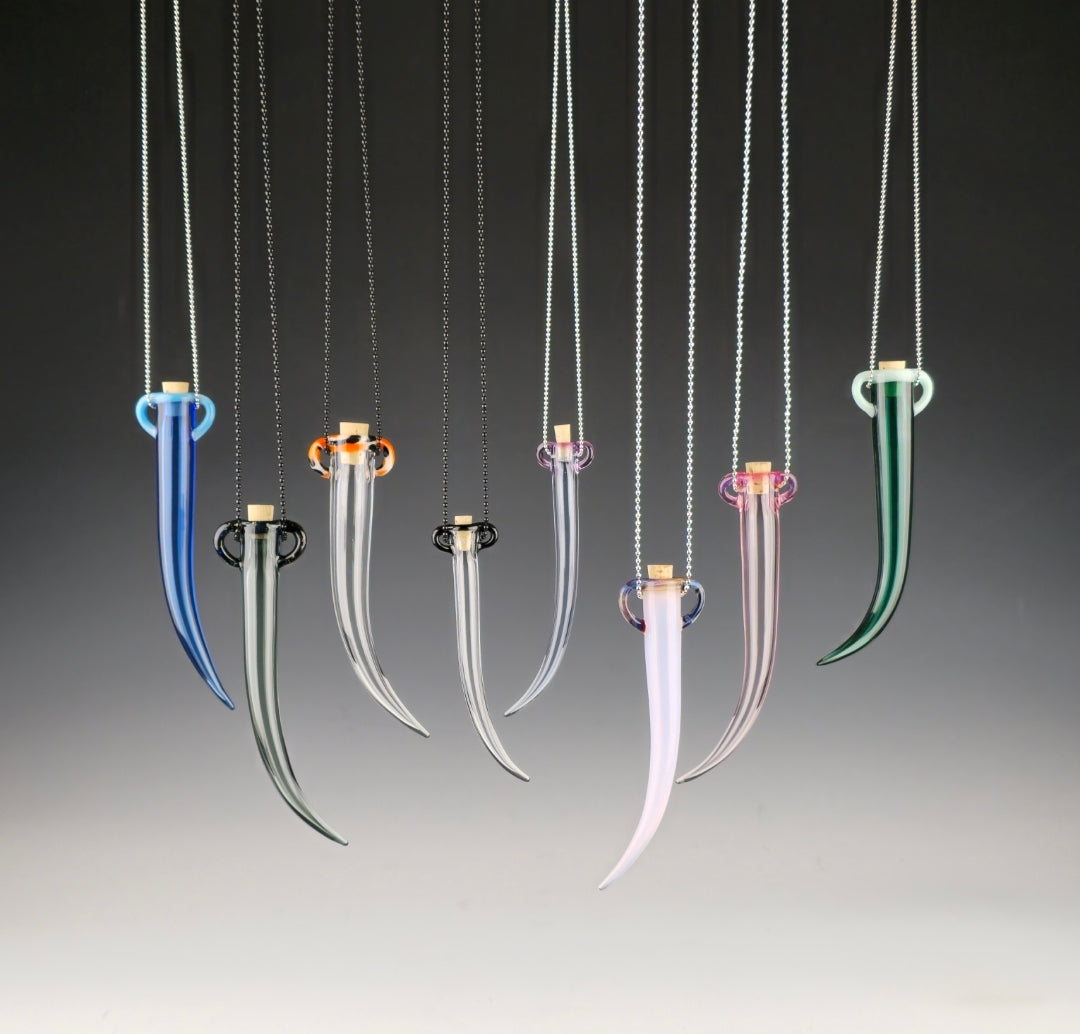 Group shot of several different colored glass whisker shaped vessels which each have 2 small handles at the top so they can be hung or worn as a necklace. A safe place to keep the fallen whiskers you find.