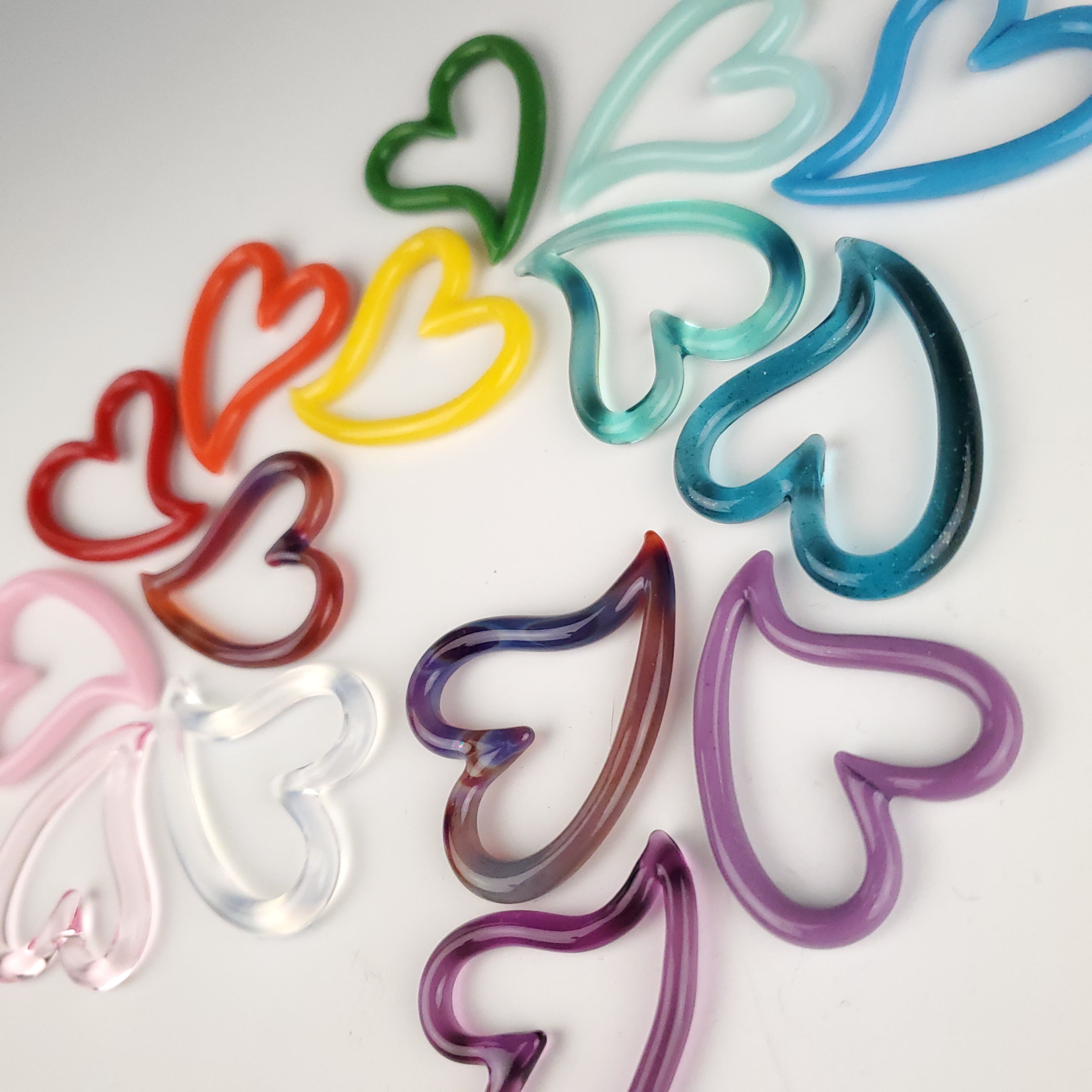 A collection of artistically arranged glass hearts in a rainbow of colors. These glass hearts are hand made necklaces and five dollars from each one sold gets donated to Mission Meow.