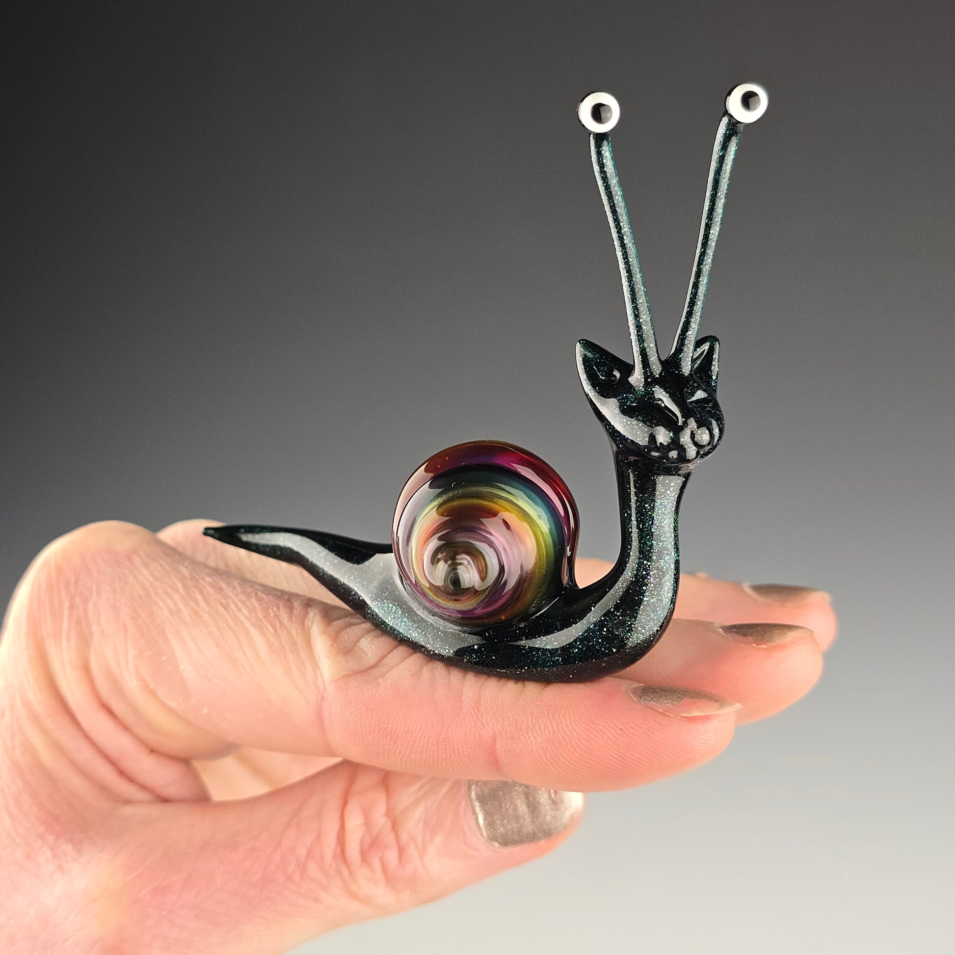 whimsical glass sculpture of a snail cat made out of sparkly dark blue glass with a metallic rainbow snail shell