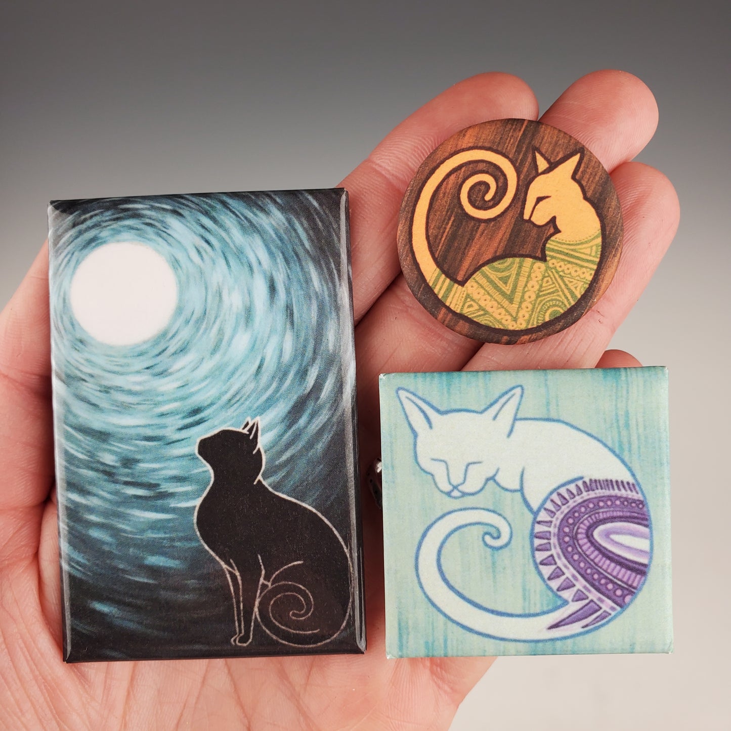 A hand holding three different magnets with artistic paintings on them. One is a silhouette of a cat, another is a cat in a yin-yang symbol, and the last one is an abstract design.