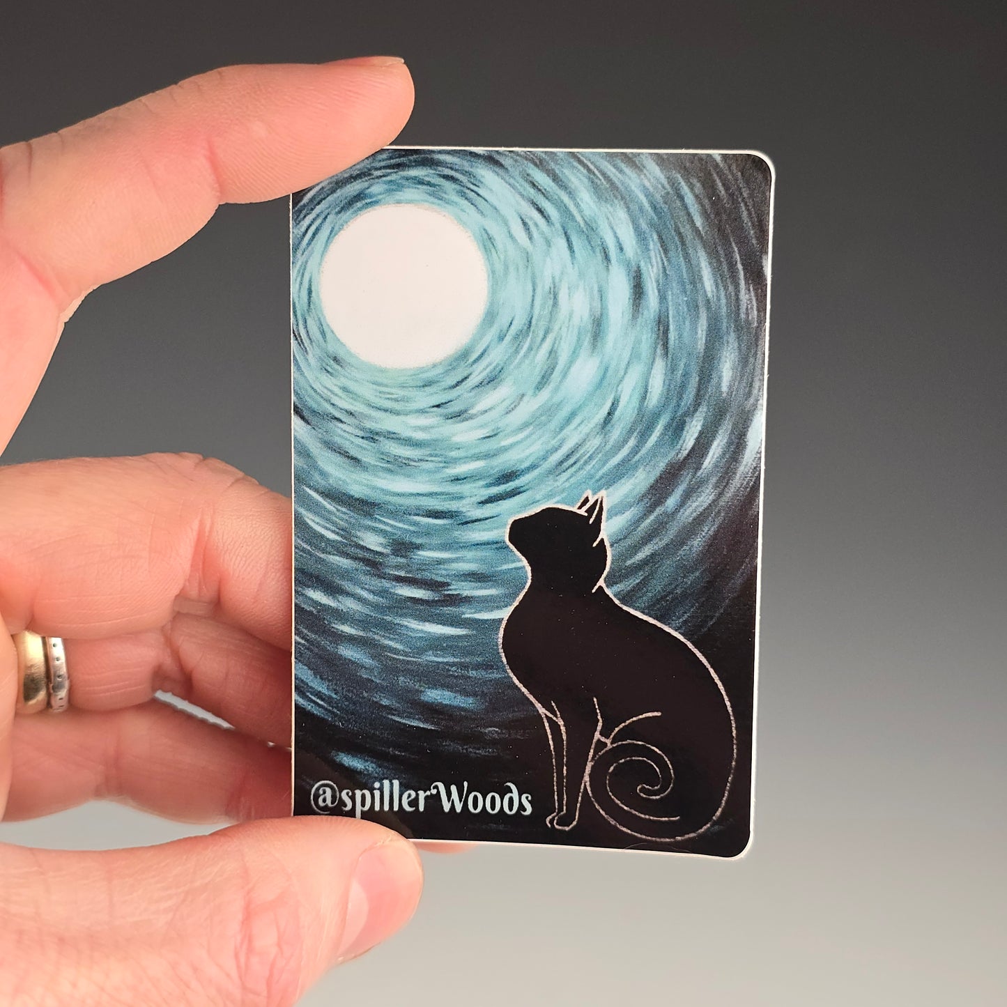 Sticker of a painting of a black cat silhouette and swirling blue and white design held by hands against a gray background