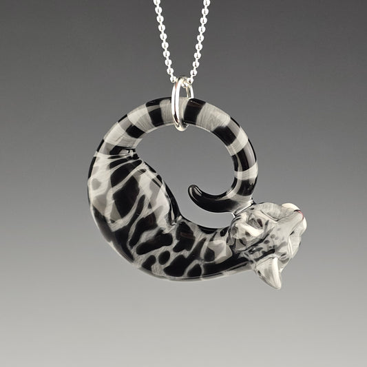 A large pendant in the shape of a sleeping tabby cat with black and gray swirls, signed and dated with a titanium pen, displayed on a ball chain.