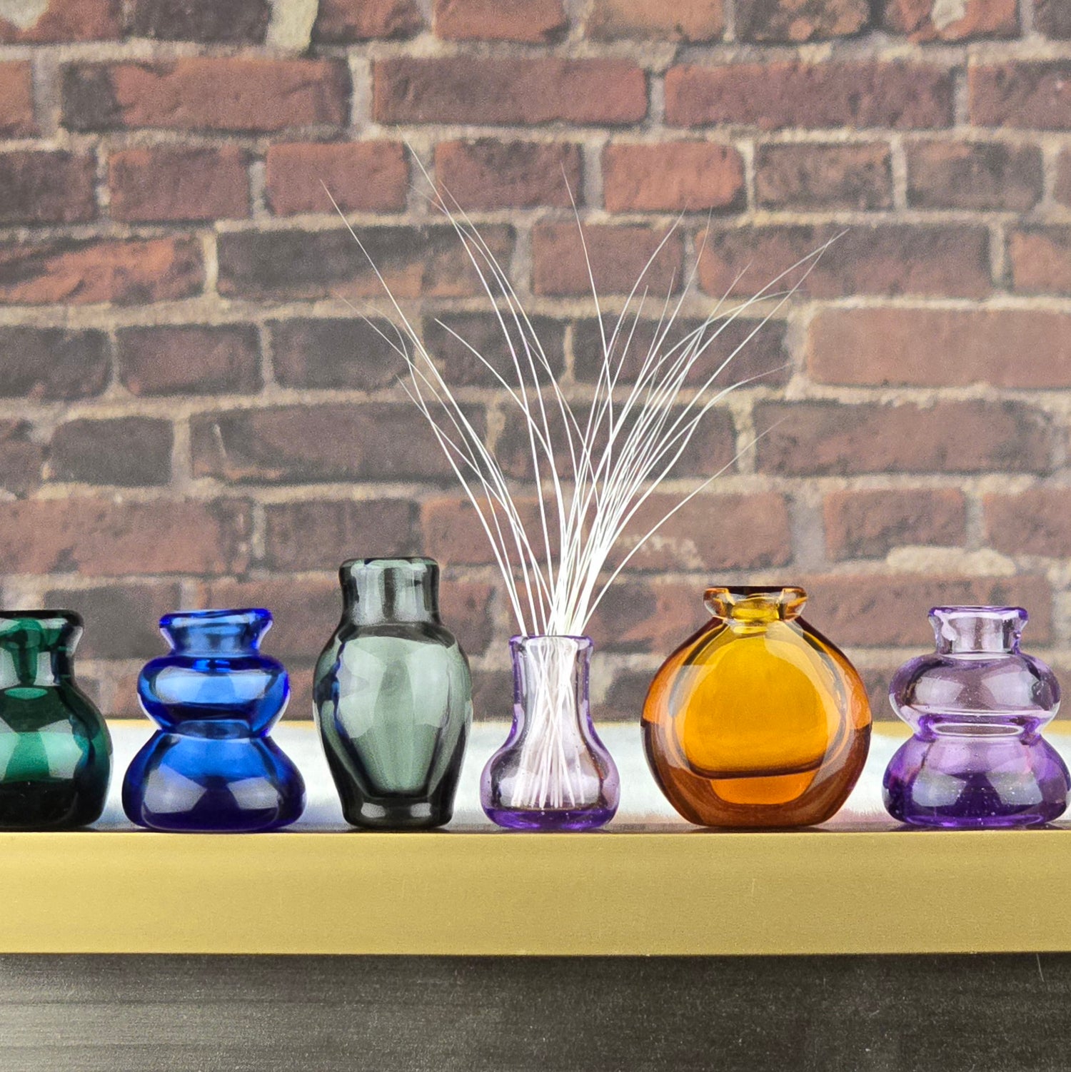 Colorful miniature glass vases filled with cat whiskers on a shelf against a brick wall